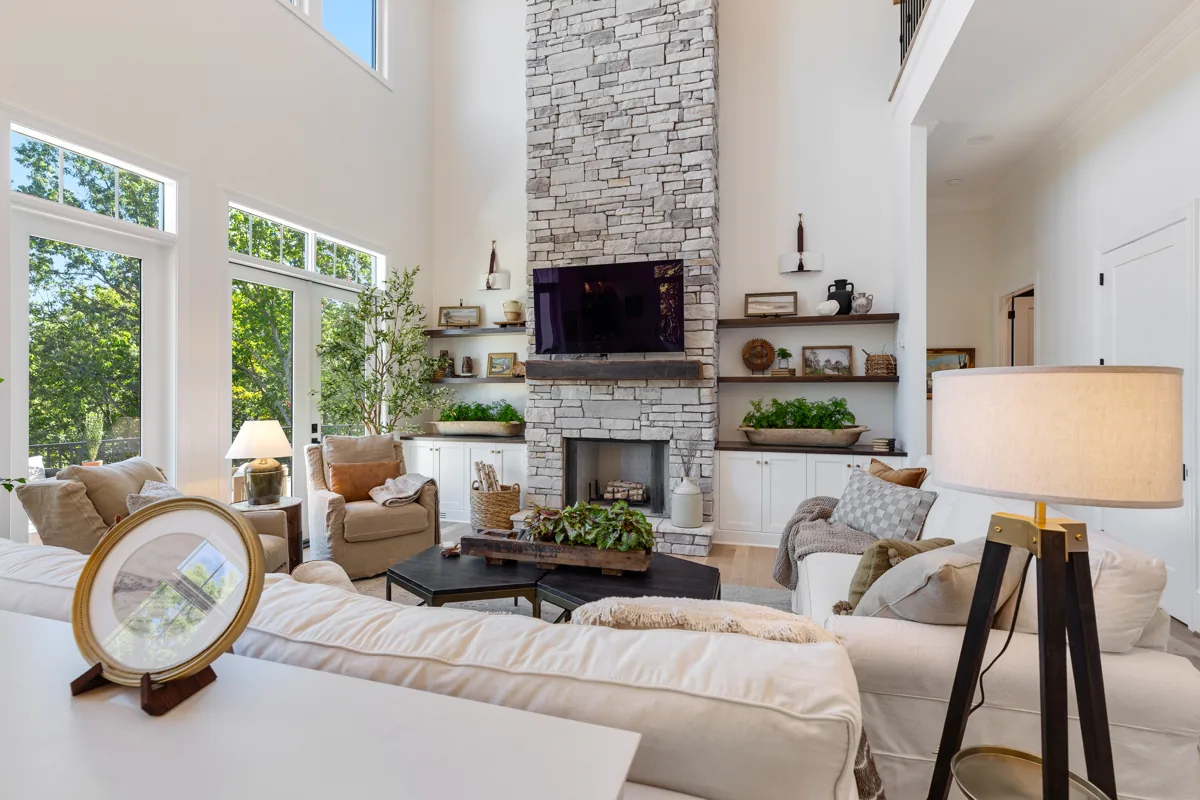 A tall stone fireplace framed by shelving, large windows, and neutral seating.