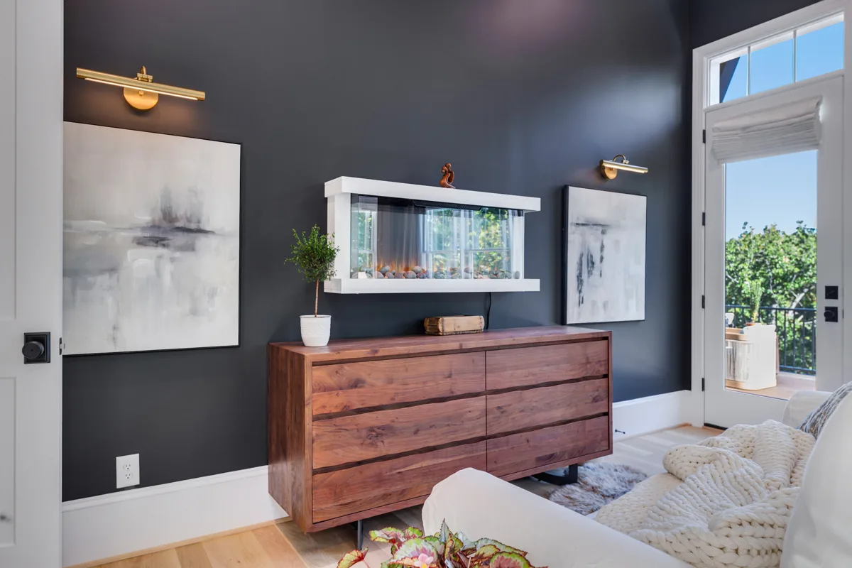 A wood chest, framed artwork, brass picture lights, and a mirror against charcoal walls.