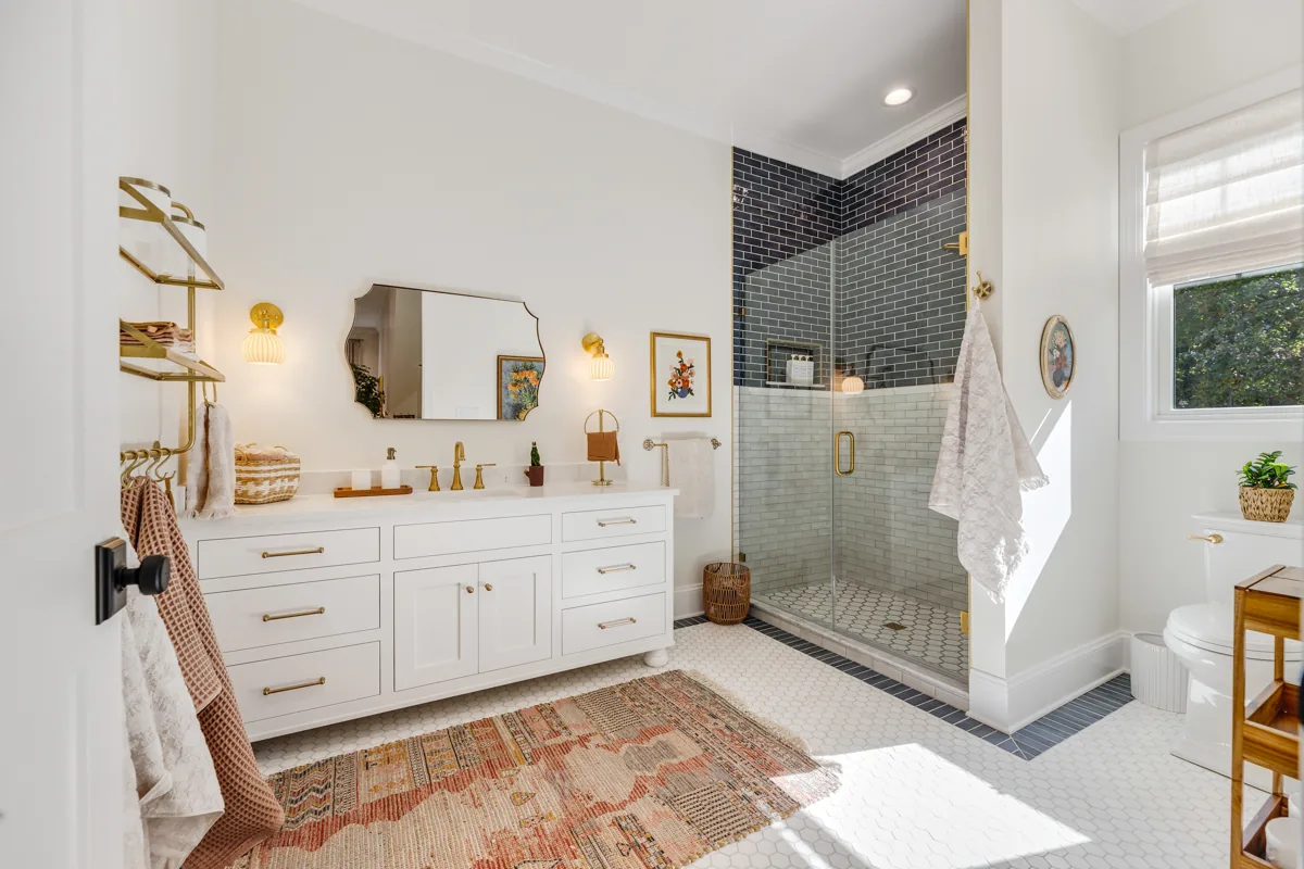 White cabinetry, gold mirrors, a vintage-style rug, and a dark tiled shower.